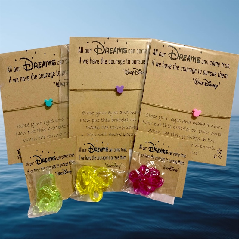 Peut inclure: Plusieurs bracelets de v&oelig;ux sur le th&egrave;me de Disney. Chaque bracelet est orn&eacute; d'un petit charme de diff&eacute;rentes couleurs, attach&eacute; &agrave; une ficelle marron et emball&eacute; sur une carte marron avec du texte. Les cartes indiquent "All our Dreams can come true..."
