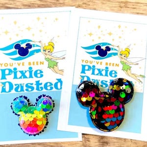 May include: Two cards with a Mickey Mouse-shaped sequin pin. The cards have the text "You've Been Pixie Dusted" with a cartoon of Tinkerbell. One pin is rainbow-colored, the other is black with colorful sequins. The background is light blue and white.