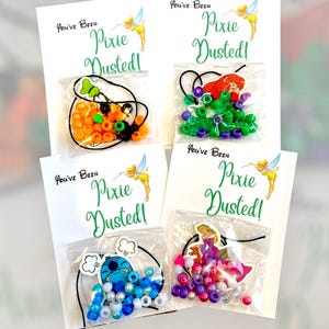 May include: Four small gift bags with the text "You've Been Pixie Dusted!" and images of Tinkerbell. Each bag contains a different beaded bracelet with a character charm, including a green and orange Halloween-themed bracelet, an Ariel bracelet, a Stitch bracelet, and a princess bracelet.