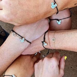 Peut inclure: Plusieurs poignets orn&eacute;s de bracelets &agrave; breloques. Certains bracelets pr&eacute;sentent un charm en forme de t&ecirc;te de Mickey Mouse, d'autres un charm circulaire dor&eacute;. Les bracelets sont sur des cha&icirc;nes noires, roses et dor&eacute;es.