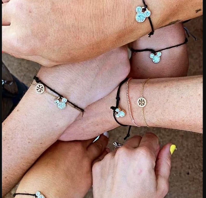 Peut inclure: Plusieurs poignets orn&eacute;s de bracelets &agrave; breloques. Certains bracelets pr&eacute;sentent un charm en forme de t&ecirc;te de Mickey Mouse, d'autres un charm circulaire dor&eacute;. Les bracelets sont sur des cha&icirc;nes noires, roses et dor&eacute;es.