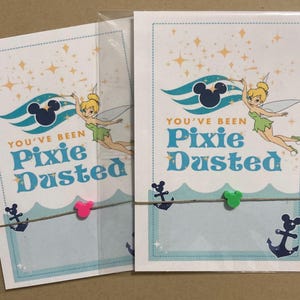 May include: Two cards with a Tinkerbell theme. Each card has the text "You've Been Pixie Dusted" with a cartoon image of Tinkerbell. The cards are decorated with a Mickey Mouse silhouette, anchors, and stars. The cards are held together with twine and small Mickey Mouse-shaped clips.