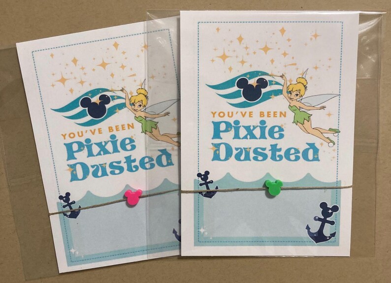May include: Two cards with a Tinkerbell theme. Each card has the text "You've Been Pixie Dusted" with a cartoon image of Tinkerbell. The cards are decorated with a Mickey Mouse silhouette, anchors, and stars. The cards are held together with twine and small Mickey Mouse-shaped clips.