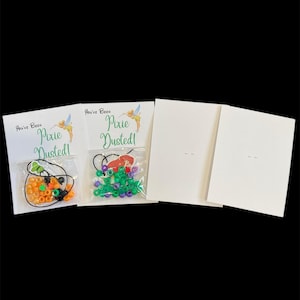 May include: Two cards with the text "You've Been Pixie Dusted!" and a Tinkerbell graphic. Two small bags of colorful beads are attached to the cards. Two blank cards are also included.