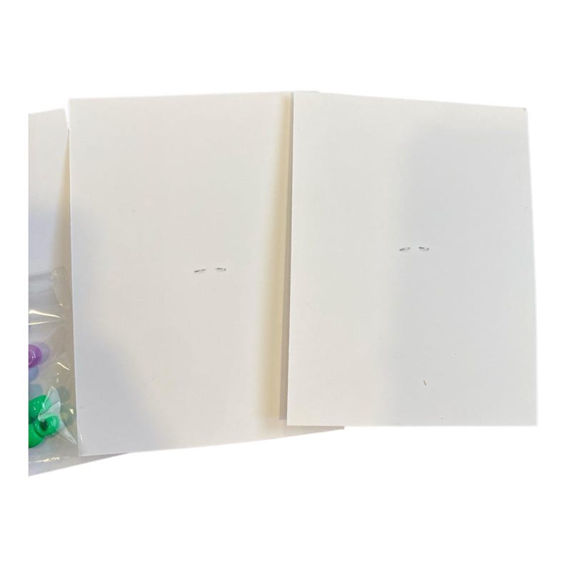 May include: A close-up shot of two white rectangular cards, possibly for crafting or stationery. The cards are clean and plain, with a few small marks. A partially visible plastic bag on the left contains colorful beads, suggesting a craft supply.