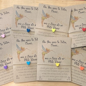 May include: Cards with the text "All You Need Is Faith, Trust, And A Little Bit Of Pixie Dust" featuring a Tinkerbell graphic. Each card has a colorful charm attached. The cards are arranged on a wooden surface.