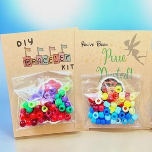 May include: Two DIY bracelet kits with colorful beads in clear plastic bags. One kit has red, purple, and green beads, and the other has blue, yellow, and red beads. The kits are on brown cardstock with the text "DIY BRACELET KIT" and "You've Been Pixie Dusted!"