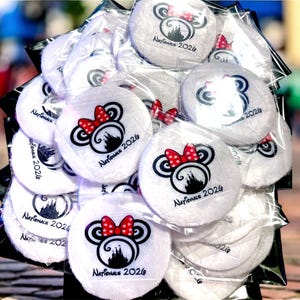 May include: A stack of white, round, plush items, each featuring a black and red Minnie Mouse design with a red bow and the text "Nationals 2026". The items are packaged in clear plastic.