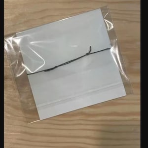 Peut inclure: Un sac en plastique transparent contenant deux cartes rectangulaires blanches et une ficelle noire. Les cartes sont empil&eacute;es et semblent vierges. Le sac est scell&eacute; et pos&eacute; sur une surface en bois brun clair.