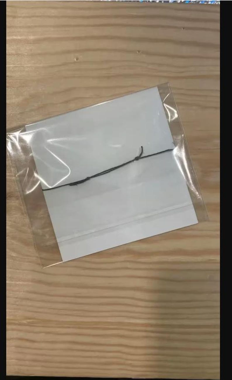 Peut inclure: Un sac en plastique transparent contenant deux cartes rectangulaires blanches et une ficelle noire. Les cartes sont empil&eacute;es et semblent vierges. Le sac est scell&eacute; et pos&eacute; sur une surface en bois brun clair.
