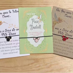 May include: Three wish bracelets with cards. The cards have text and images of Tinkerbell. The bracelets have a Mickey Mouse charm. The cards are white, green, and brown. The text on the cards reads "All you need is faith, trust... and a little bit of pixie dust."