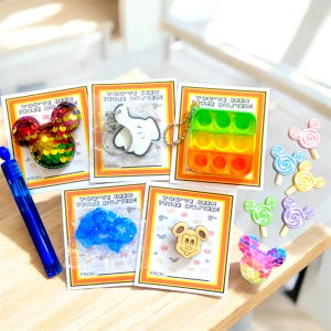 May include: A collection of Disney-themed gifts and favors. Includes Mickey Mouse-shaped items like a sequined head, keychains, and lollipops. Cards with the text "You've Been Pixie Dusted!" are also present, along with a blue bubble wand.