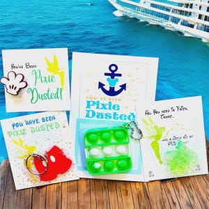 May include: A collection of themed items with the phrase "Pixie Dusted" on cards and a pop-it toy. The items include a Mickey Mouse hand-shaped magnet, a keychain, and a green and white pop-it toy. The background shows a cruise ship and ocean.
