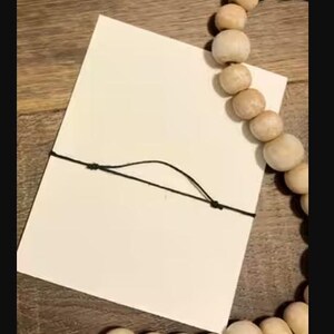 Peut inclure: Une carte blanche vierge attach&eacute;e avec une ficelle noire, &agrave; c&ocirc;t&eacute; d'un chapelet de perles en bois de couleur claire. Les perles sont rondes et ont une finition naturelle. La carte est rectangulaire et semble &ecirc;tre en papier.