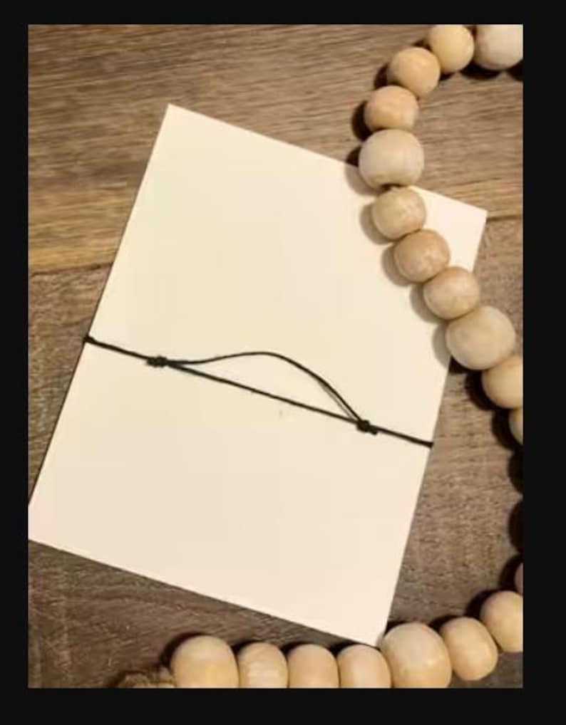 Peut inclure: Une carte blanche vierge attach&eacute;e avec une ficelle noire, &agrave; c&ocirc;t&eacute; d'un chapelet de perles en bois de couleur claire. Les perles sont rondes et ont une finition naturelle. La carte est rectangulaire et semble &ecirc;tre en papier.