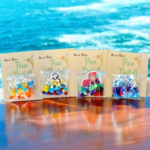 May include: Four small clear bags filled with colorful beads, each paired with a card that reads "You've Been Pixie Dusted!". The beads are in various colors, including orange, blue, yellow, and purple. The background is a blurred view of the ocean.