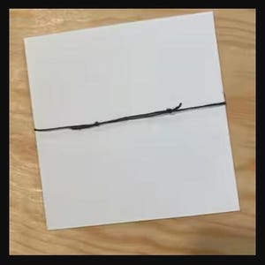 Peut inclure: Une carte carr&eacute;e blanche attach&eacute;e avec une ficelle noire. La carte est centr&eacute;e dans le cadre et la ficelle est nou&eacute;e. Le fond est une surface en bois brun clair.