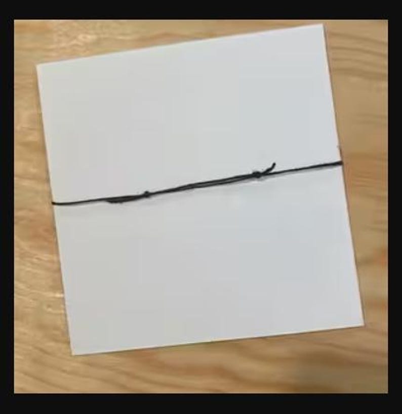 Peut inclure: Une carte carr&eacute;e blanche attach&eacute;e avec une ficelle noire. La carte est centr&eacute;e dans le cadre et la ficelle est nou&eacute;e. Le fond est une surface en bois brun clair.