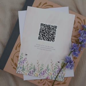 K&ouml;nnte beinhalten: Briefpapier mit floralem Design und QR-Code, auf einem rosa und grauen Buch. Das Briefpapier hat einen Aquarell-Blumenrand mit Bienen und den Text "PLEASE SCAN THE QR CODE OR VISIT WEDDINGWEBSITE.COM TO VIEW OUR REGISTRY."