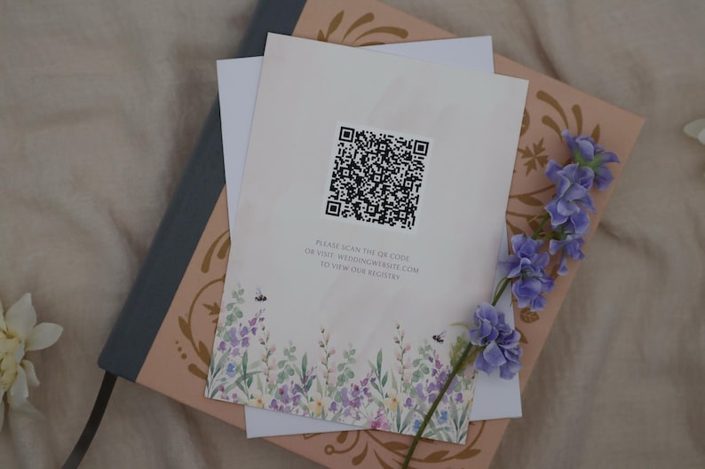 K&ouml;nnte beinhalten: Briefpapier mit floralem Design und QR-Code, auf einem rosa und grauen Buch. Das Briefpapier hat einen Aquarell-Blumenrand mit Bienen und den Text "PLEASE SCAN THE QR CODE OR VISIT WEDDINGWEBSITE.COM TO VIEW OUR REGISTRY."