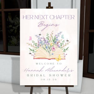 May include: A welcome sign on an easel with the text "Her Next Chapter Begins." The sign features an open book with floral and butterfly illustrations. Below, it reads "Welcome to Hannah Alexander's Bridal Shower 08.15.20."