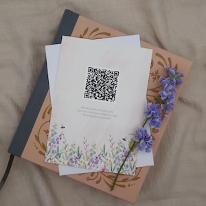 K&ouml;nnte beinhalten: Briefpapier liegt auf einem dekorativen Buch. Die oberste Karte hat einen QR-Code und ein Blumenmuster. Ein Zweig lila Blumen und zwei wei&szlig;e Blumen sind auf einer neutralen Oberfl&auml;che angeordnet.