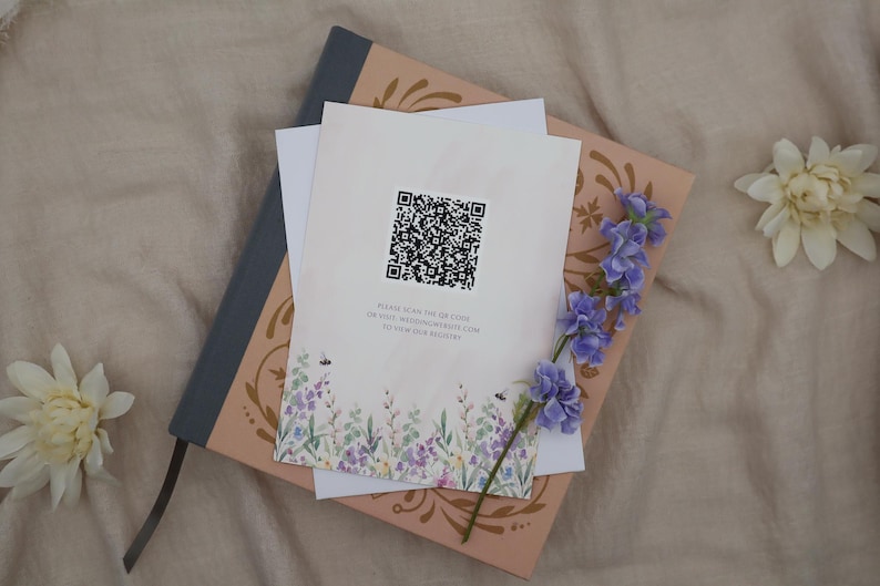 K&ouml;nnte beinhalten: Briefpapier liegt auf einem dekorativen Buch. Die oberste Karte hat einen QR-Code und ein Blumenmuster. Ein Zweig lila Blumen und zwei wei&szlig;e Blumen sind auf einer neutralen Oberfl&auml;che angeordnet.
