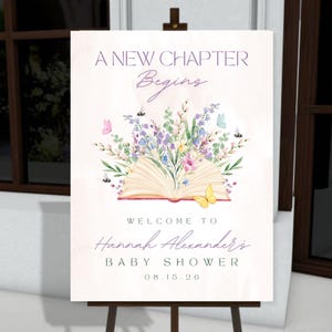 May include: A sign on an easel with the text "A New Chapter Begins" and "Welcome to Hannah Alexander's Baby Shower 08.15.20". The sign features a watercolor illustration of an open book with flowers and butterflies.