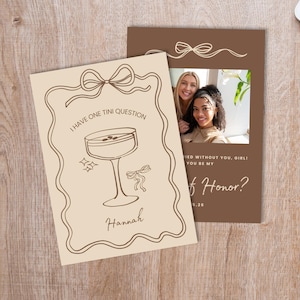 May include: Two invitation cards on a wooden surface. One card is beige with a cocktail illustration and the text "I HAVE ONE TINI QUESTION" and "Hannah". The other card is brown with a photo and the text "Will you be my Maid of Honor?"