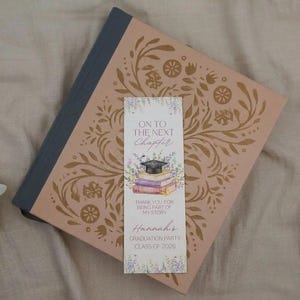 Puede incluir: Un marcap&aacute;ginas con tem&aacute;tica de graduaci&oacute;n descansa sobre un libro decorativo con un dise&ntilde;o floral. El marcap&aacute;ginas dice "On to the Next Chapter" e incluye una ilustraci&oacute;n de un birrete. El libro es de color melocot&oacute;n y tiene una espina dorsal azul oscuro.