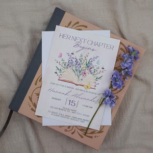 K&ouml;nnte beinhalten: Eine Einladung zur Brautparty mit dem Text "HER NEXT CHAPTER Begins" und Details f&uuml;r Hannah Alexanders Party. Die Einladung zeigt Blumen- und Schmetterlingsillustrationen mit einem Blau- und Rosafarbschema. Ein blaues Notizbuch und Blumen sind ebenfalls vorhanden.