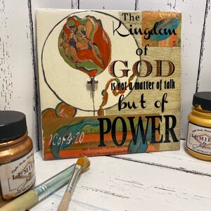 May include: A wooden sign with a gold background and a floral design. The sign reads "The Kingdom of God is not a matter of talk but of power" and includes the scripture reference "1 Cor 4:20".