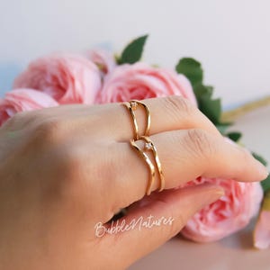 May include: Two gold-toned rings with a wavy, open-gap design, each featuring a single small, clear, round-cut stone set at the top. The rings have a polished finish and are worn on two fingers. The text BubbleNatures is visible in white script.