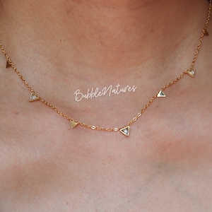 Puede incluir: Un collar de cadena dorada con pequeños colgantes triangulares, cada uno con un cristal transparente en el centro. El collar está en el cuello de una modelo.
