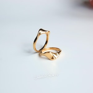 May include: Two gold rings with a fluid, organic design. One ring has a wave-like form, the other a signet style with a curved surface. The rings are displayed on a white surface, reflecting light and highlighting their polished finish. The rings are made of gold.