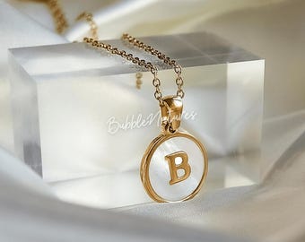 May include: A gold-colored necklace with a pendant featuring the letter "B" on a white background. The pendant is round and framed in gold. The necklace is displayed on a clear, rectangular block.