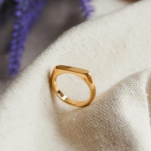Pode incluir: Um anel dourado com uma parte superior retangular, colocado sobre um tecido branco. O design do anel é simples e moderno. Flores roxas são visíveis no fundo. Uma joia.