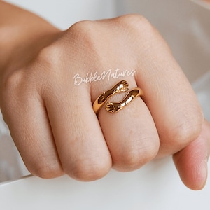 Könnte beinhalten: Ein goldfarbener Ring in Form von zwei sich umarmenden Händen. Der Ring wird an einem Finger getragen, wobei sich die Hände um den Finger legen. Der Ring hat eine glatte, polierte Oberfläche vor einem neutralen Hintergrund. Der Ring ist ein Schmuckstück.