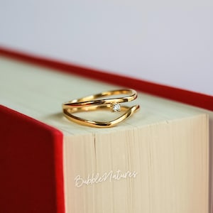 Puede incluir: Un anillo dorado con una pequeña gema transparente. El anillo tiene un diseño abierto único con dos bandas curvas. Descansa sobre un libro con una cubierta roja. El texto "BubbleNatures" es visible en el libro.