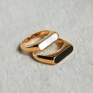 May include: Two gold rings with rectangular inlays. The top ring has a white inlay and the bottom ring has a black inlay.