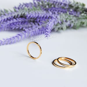 Puede incluir: Tres anillos de banda delgada de color dorado se muestran sobre una superficie blanca, con una ramita de lavanda púrpura en el fondo. Los anillos son simples y elegantes, con un diseño minimalista. El nombre de la marca "BubbleNatures" está escrito en cursiva.