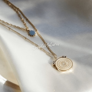 Op de afbeelding: Goudkleurige ketting met meerdere lagen, met een zonnestraal hanger en een ronde hanger met een oogontwerp. De ketting ligt op een witte satijnen ondergrond. De merknaam "Bubble Natures" is zichtbaar.