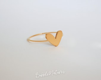 May include: A gold-colored ring featuring a heart-shaped design. The ring has a thin band and a solid heart shape. The ring is displayed on a white surface. The brand name "BubbleNatures" is written in cursive on the surface.