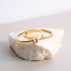 Puede incluir: Dos anillos de oro, uno con una piedra clara de corte cuadrado y el otro con pequeñas piedras claras, descansan sobre una superficie blanca y texturizada. Los anillos se exhiben sobre una piedra blanca sobre un fondo suave y neutro.