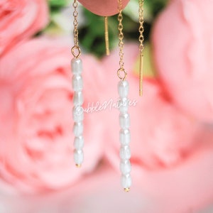 Op de afbeelding: Een paar gouden kettingoorbellen met witte parels die eraan hangen. De oorbellen zijn tegen een roze achtergrond.