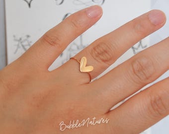 May include: A gold-colored ring featuring a heart-shaped design. The ring is worn on a finger, with the heart prominently displayed. The background includes a greeting card with the word "Birthday" and the text "BubbleNatures".