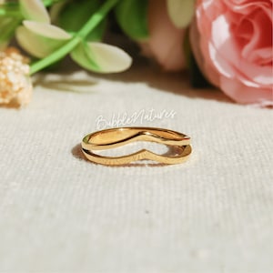 Op de afbeelding: Een goudkleurige ring met een dubbele band. De ring heeft een V-vormige bovenband en een rechte onderste band. De ring staat tegen een zachte, neutrale achtergrond met bloemaccenten. De tekst "BubbleNatures" is zichtbaar.