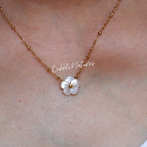 May include: A delicate gold-toned necklace featuring a small, white flower pendant with a gold center. The chain is adorned with small gold beads. The necklace is displayed on a person's skin. The text "BubbleNatures" is visible.
