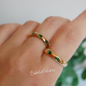 May include: Two gold rings, each set with emerald green gemstones. The rings are on a hand, with the text "BubbleNatures" in cursive script at the bottom. The rings have a smooth, polished finish against a soft, neutral background.