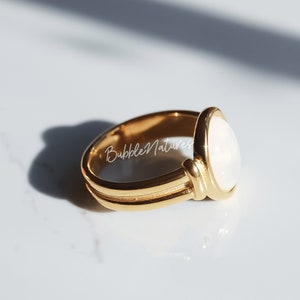 May include: A gold ring featuring a white oval gemstone. The ring has a double band and a textured design.